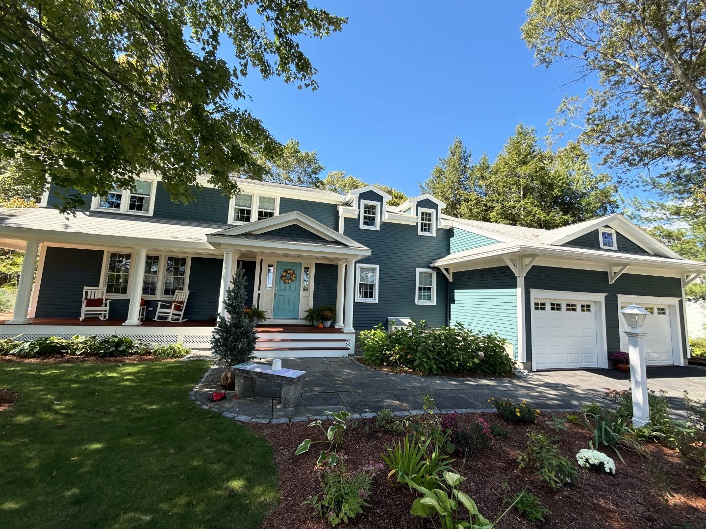 A two-story blue house with white trim and a covered porch featuring rocking chairs, a teal front door, and an attached two-bay garage. A stone path and landscaped garden beds sit in front, with trees and a blue sky in the background.