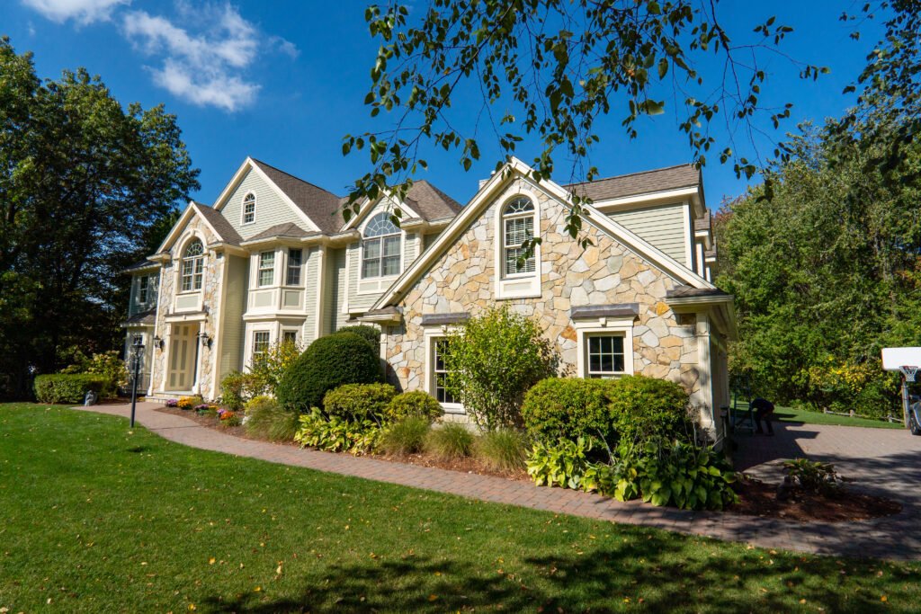 Exterior Painters Near Me in Newton, MA: A Homeowner’s Guide to Hiring the Right Painter 6 Angled exterior view of a large two-story home with light siding and prominent stone accents, arched windows, and a dark shingled roof. A brick walkway borders landscaped shrubs and a green lawn, with mature trees and a clear blue sky framing the property.