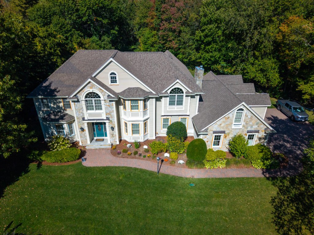 What Factors Affect Exterior Painting Costs in Massachusetts? (2026 Guide) 13 Aerial view of a large two-story suburban home with light gray siding, stone accents, and a dark shingled roof, surrounded by mature trees and landscaped shrubs. A curved brick walkway leads to the front entrance, and a driveway with a parked truck is visible on the right side of the property.