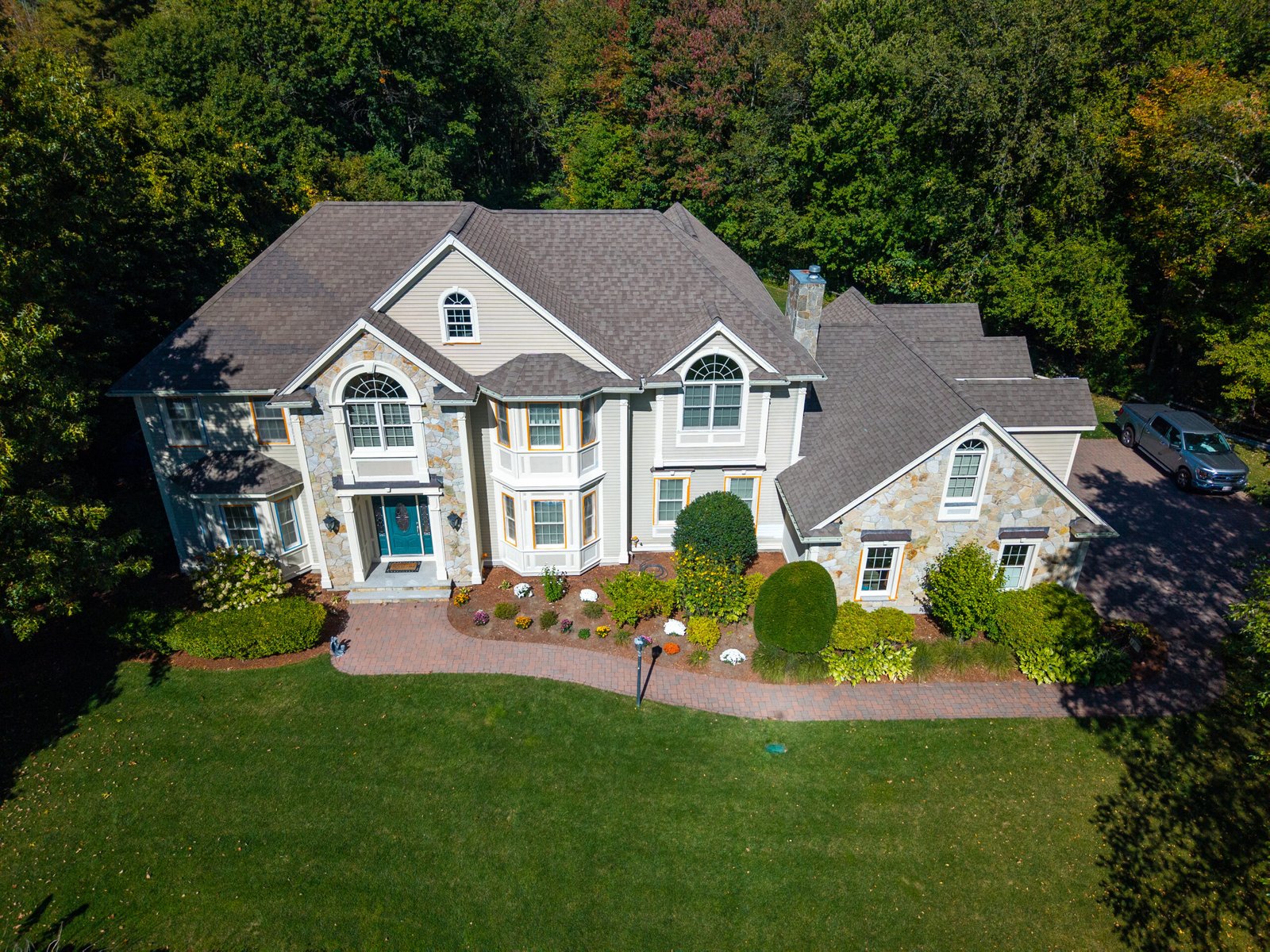 Aerial view of a large two-story suburban home with light gray siding, stone accents, and a dark shingled roof, surrounded by mature trees and landscaped shrubs. A curved brick walkway leads to the front entrance, and a driveway with a parked truck is visible on the right side of the property.