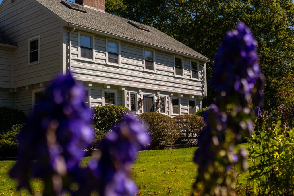 How to Choose the Best Residential Painting Company for Your Home 7 Ground-level view of a two-story home with light gray horizontal siding and white trim, partially framed by out-of-focus purple flowers in the foreground. Sunlight highlights the manicured lawn, shrubs, and the home’s symmetrical windows, with tall trees in the background.