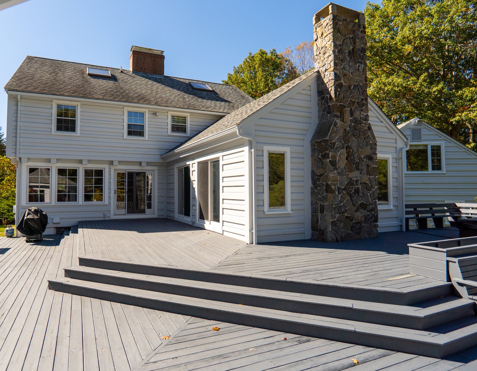 Rear exterior view of a light gray two-story home with white trim, a large raised wooden deck, and a prominent stone chimney. Sliding glass doors and windows open onto the deck, with trees and a clear sky in the background.